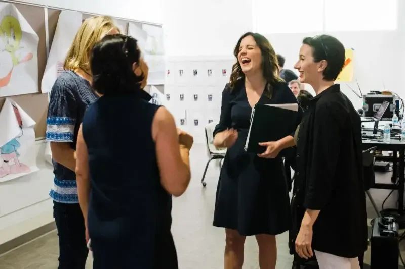 Cuatro personas de pie conversando en una sala de trabajo; una de ellas se ríe