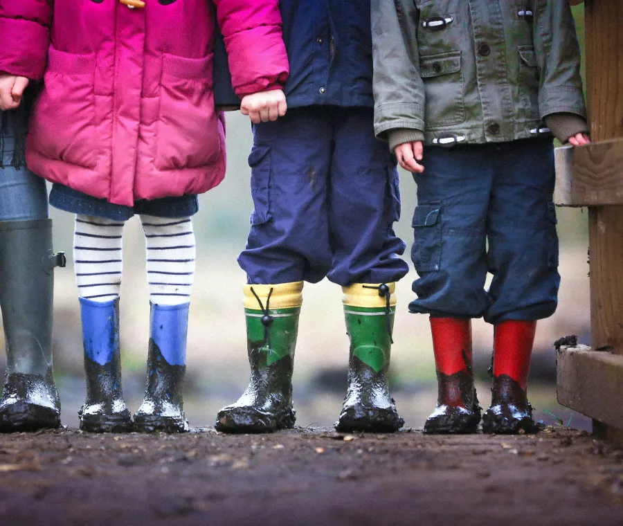 Four children in muddy rain boots standing side by side on a wooden walkway.